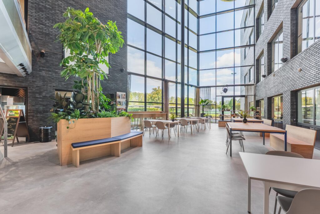 Moderne, lichte lobby of kantineruimte aan de Prof. W.H. Keesomlaan met hoge ramen, plantenbakken en zitgelegenheid.